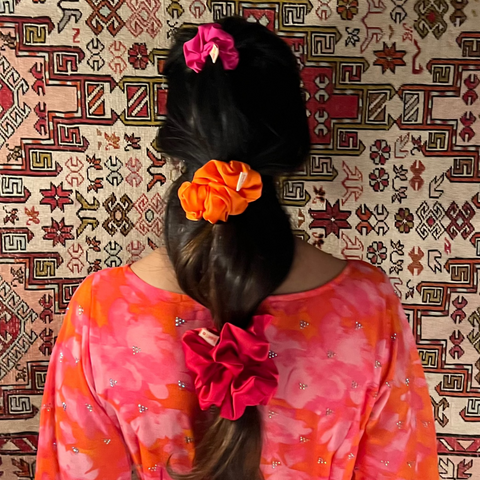 Person wearing a pink and orange patterned dress with hair tied back using colorful hair accessories on a patterned rug.