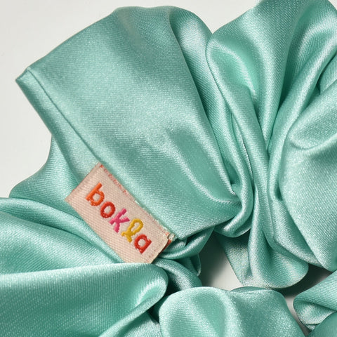 Turquoise satin scrunchie with a brand label on a light gray background